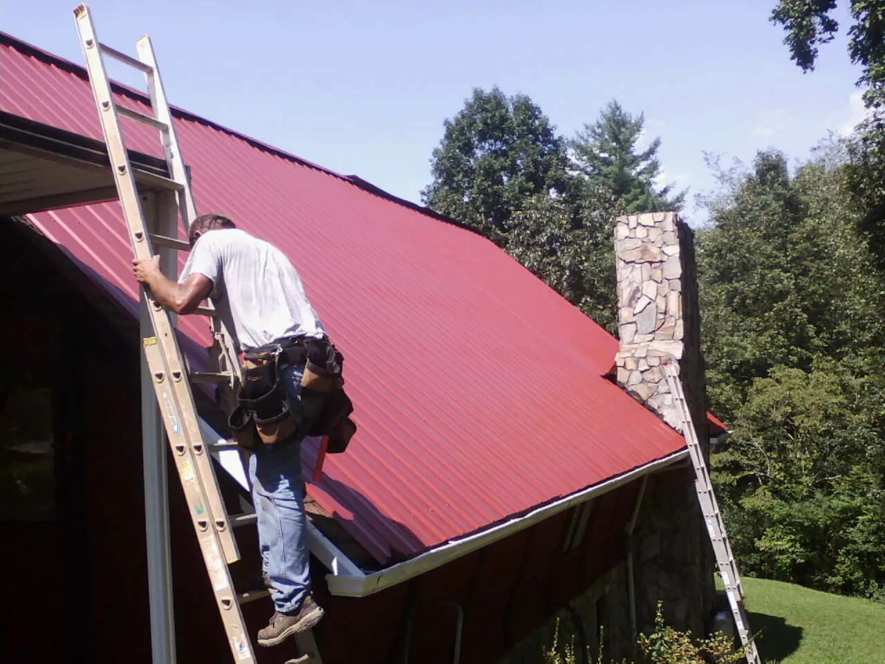 Roofer on a ladder beside a red metal roof and stone chimney — Soffit & Fascia Repair by Roofing Brothers in Cedar Grove